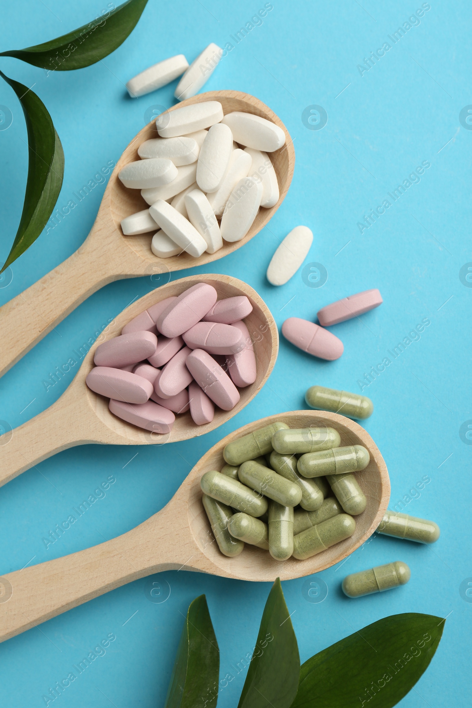 Photo of Different vitamin pills in spoons and green leaves on light blue background, flat lay