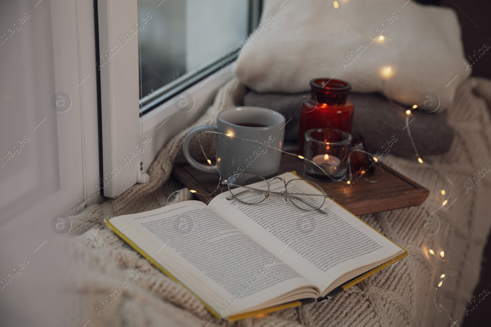 Photo of Cup of hot drink, open book and Christmas lights on knitted blanket near window. Cozy season