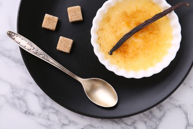 Photo of Delicious creme brulee in bowl, vanilla pod, sugar cubes and spoon on white marble table, top view
