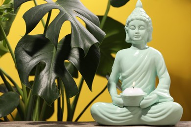 Buddhism religion. Decorative Buddha statue with burning candle on wooden table and monstera against yellow wall