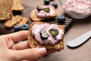 Photo of Woman holding tasty sandwich with cream cheese and blueberries at wooden table, closeup