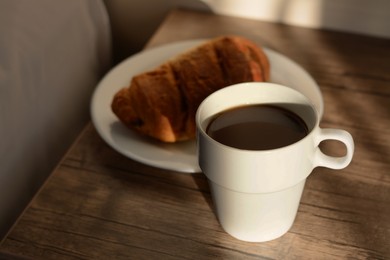Cup of coffee and croissant on wooden night stand in morning