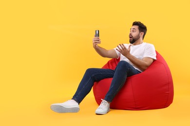 Shocked young man using smartphone on bean bag chair against yellow background, space for text