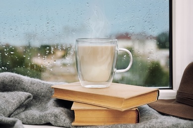 Image of Cup of delicious hot drink, books and scarf on window sill after rain 