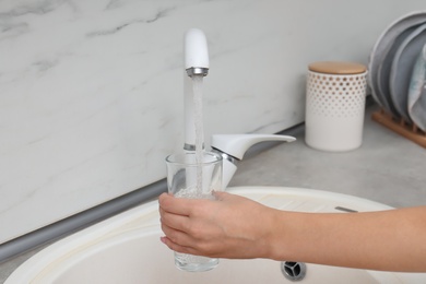 Woman filling glass with water from faucet in kitchen, closeup