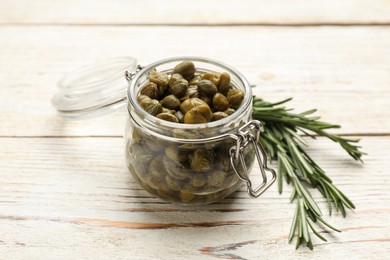 Photo of Tasty capers in glass jar and rosemary on white wooden table