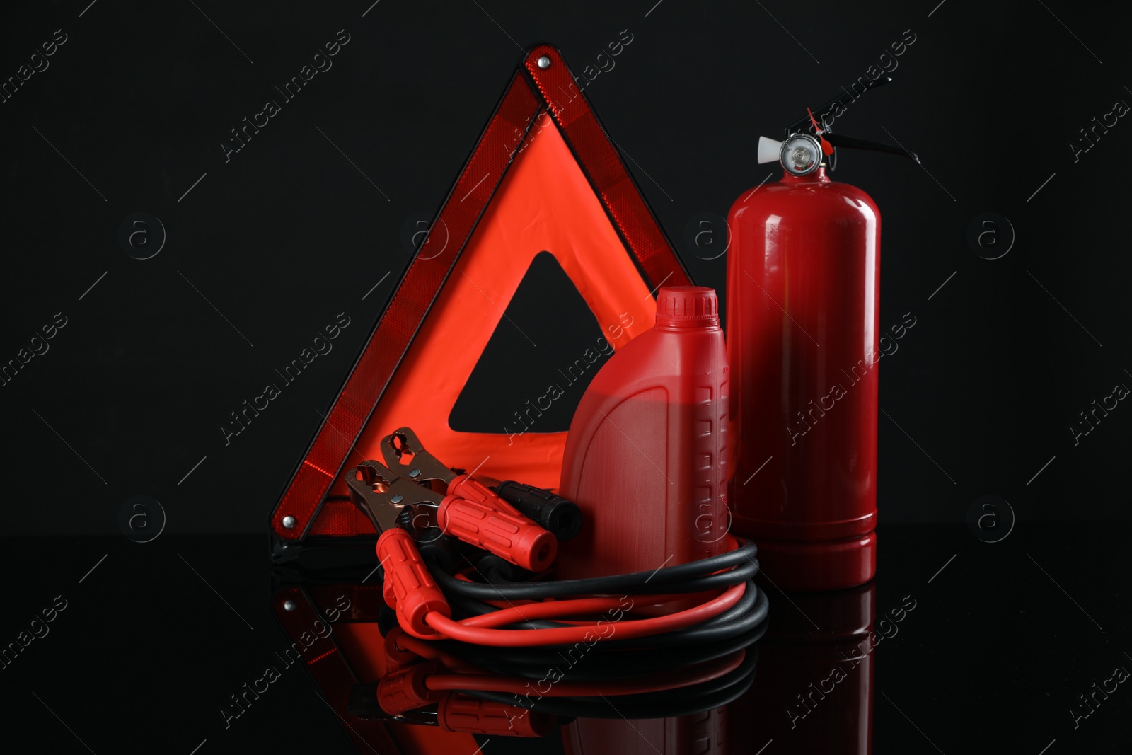 Photo of Emergency warning triangle, red fire extinguisher, battery jumper cables and motor oil on black background. Car safety