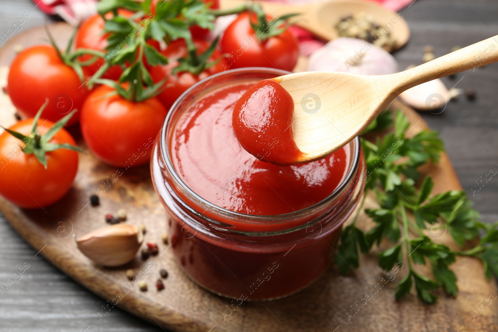 Photo of Jar and spoon with tasty ketchup, fresh tomatoes, parsley and spices on grey wooden table, closeup