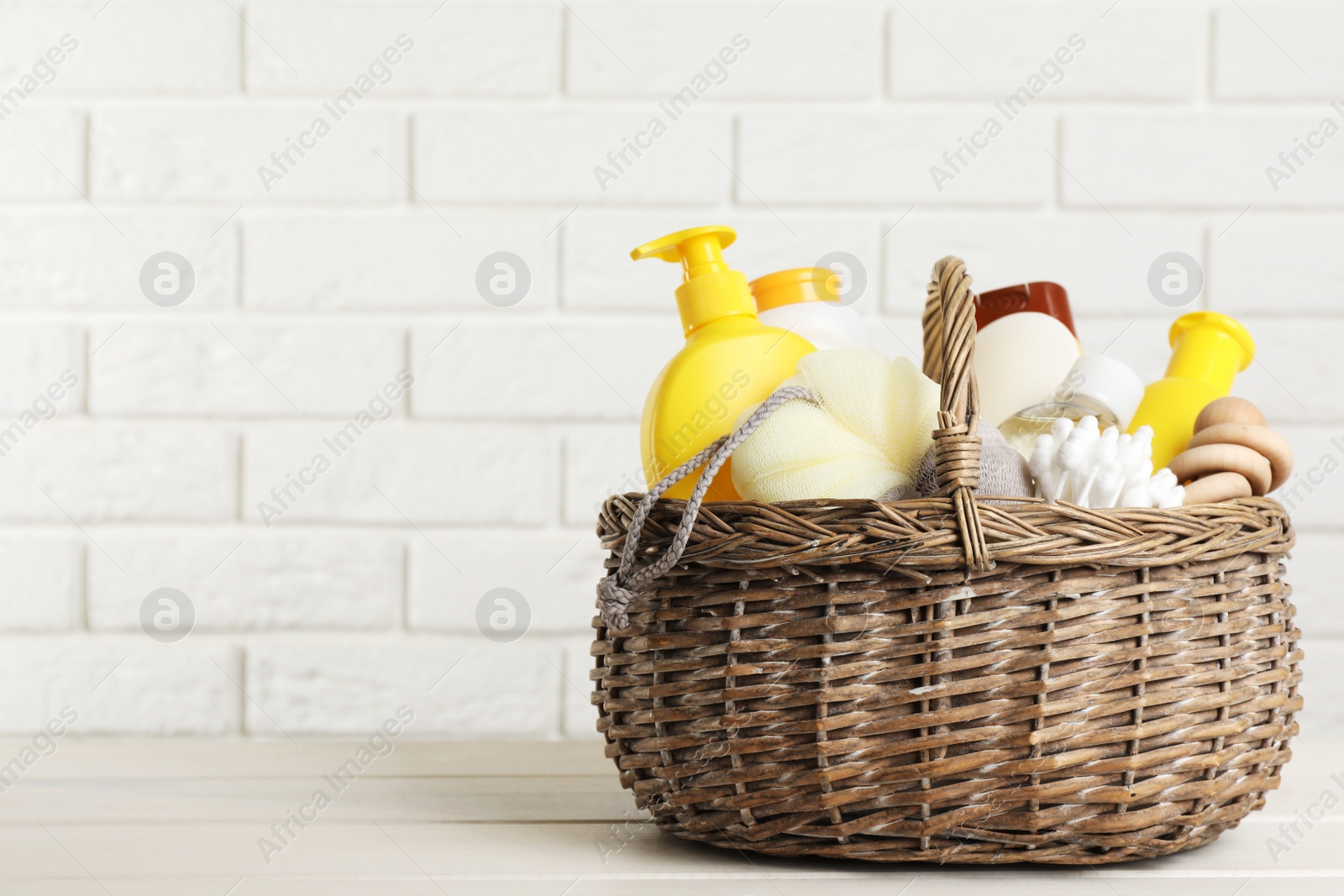 Photo of Wicker basket full of different baby cosmetic products, accessories and toy on white wooden table. Space for text