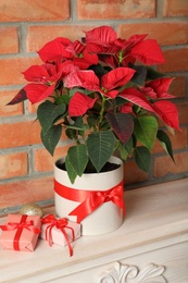 Photo of Beautiful poinsettia (traditional Christmas flower) and gifts on chest of drawers near brick wall