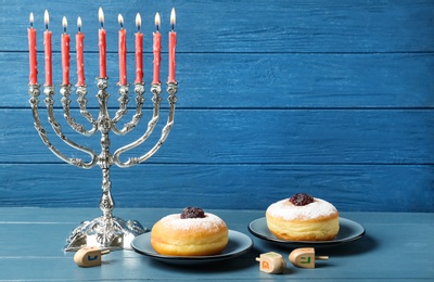 Photo of Silver menorah, dreidels with He, Pe, Nun, Gimel letters and sufganiyot on blue wooden table, space for text. Hanukkah symbols