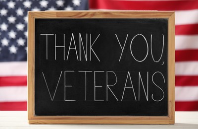 Photo of Blackboard with phrase Thank You, Veterans on white wooden table against American flag. Memorial Day