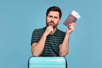 Thoughtful man with passport, suitcase and tickets on light blue background