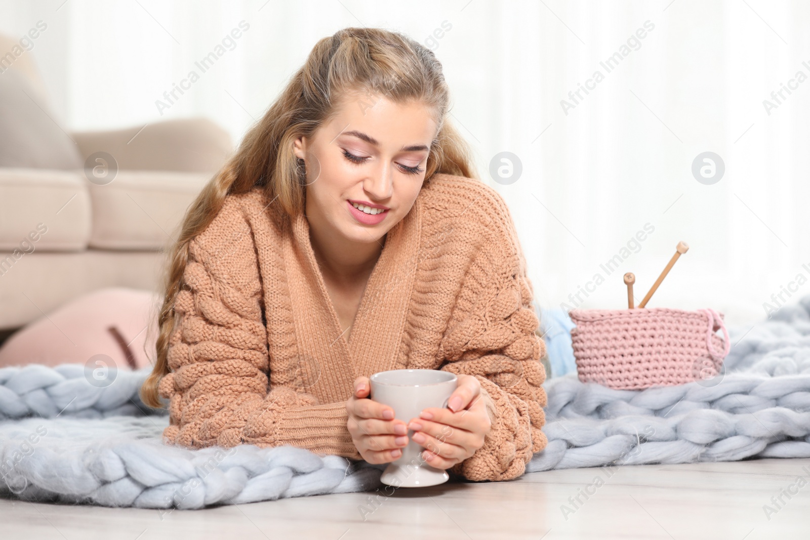Photo of Attractive smiling young woman in cozy warm sweater with cup of hot drink lying on floor at home. Space for text
