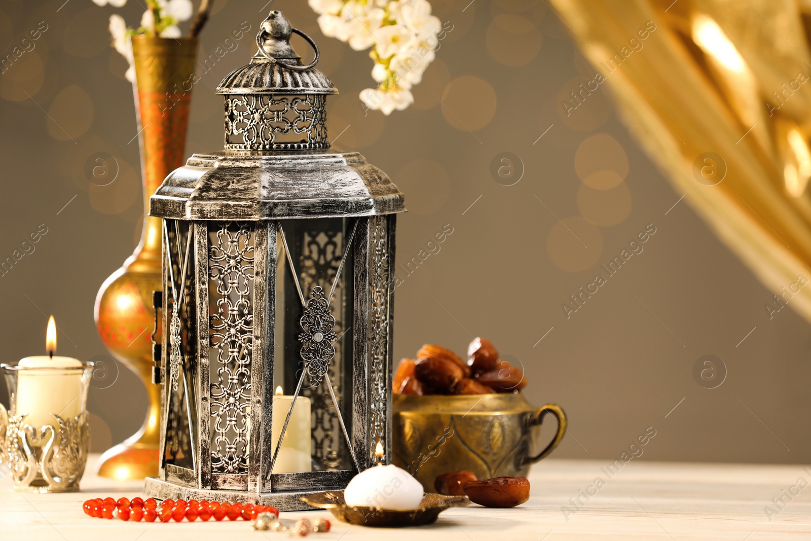 Photo of Arabic lantern, misbaha, candles, dates and flowers on table against blurred lights, space for text