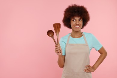 Photo of Happy young woman in apron holding spoon and spatula on pink background