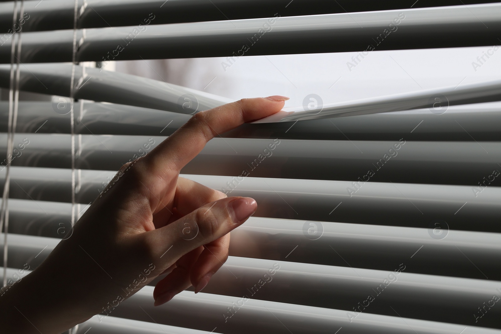 Photo of Woman separating slats of white blinds indoors, closeup