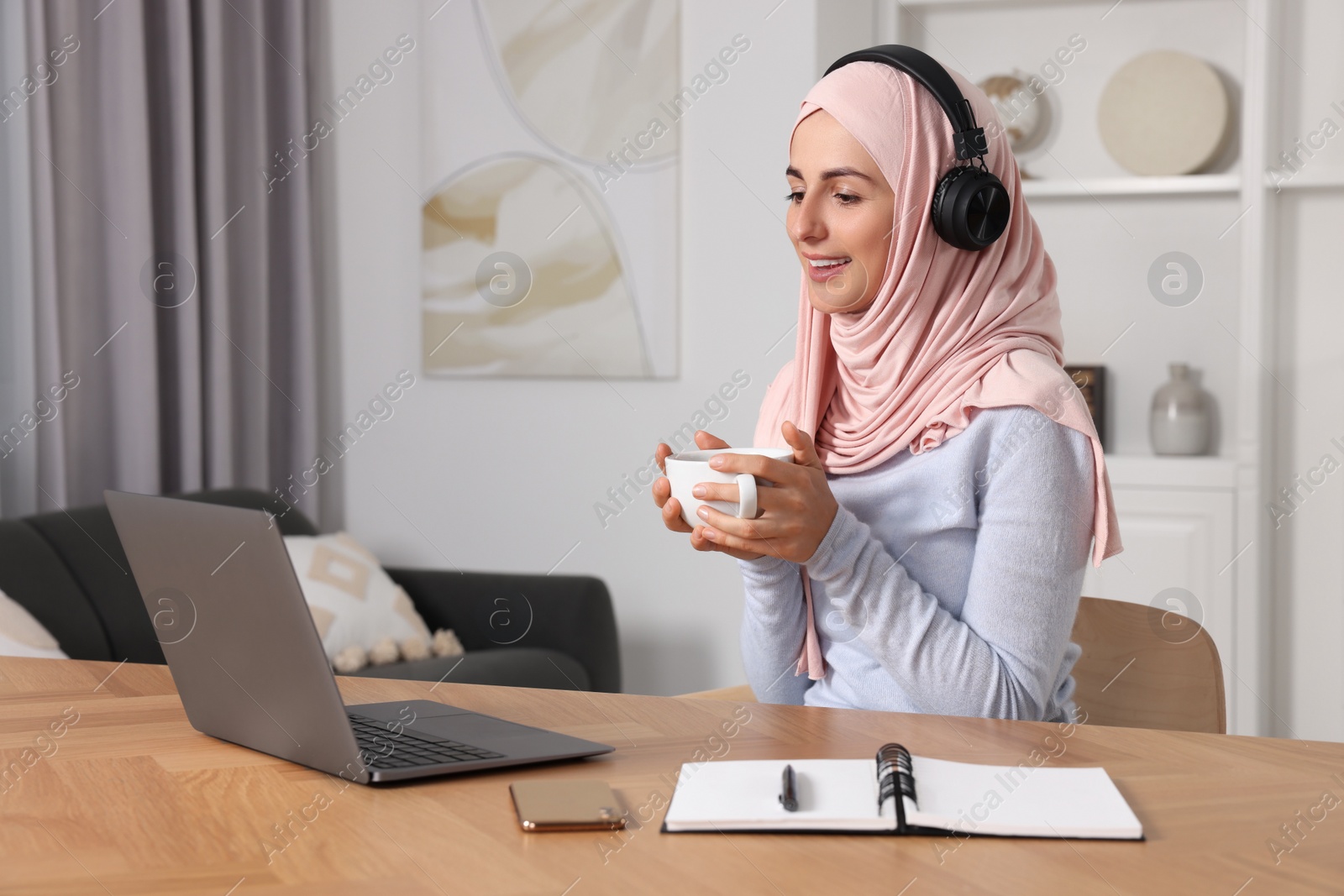 Photo of Muslim woman with cup of coffee using laptop at wooden table in room