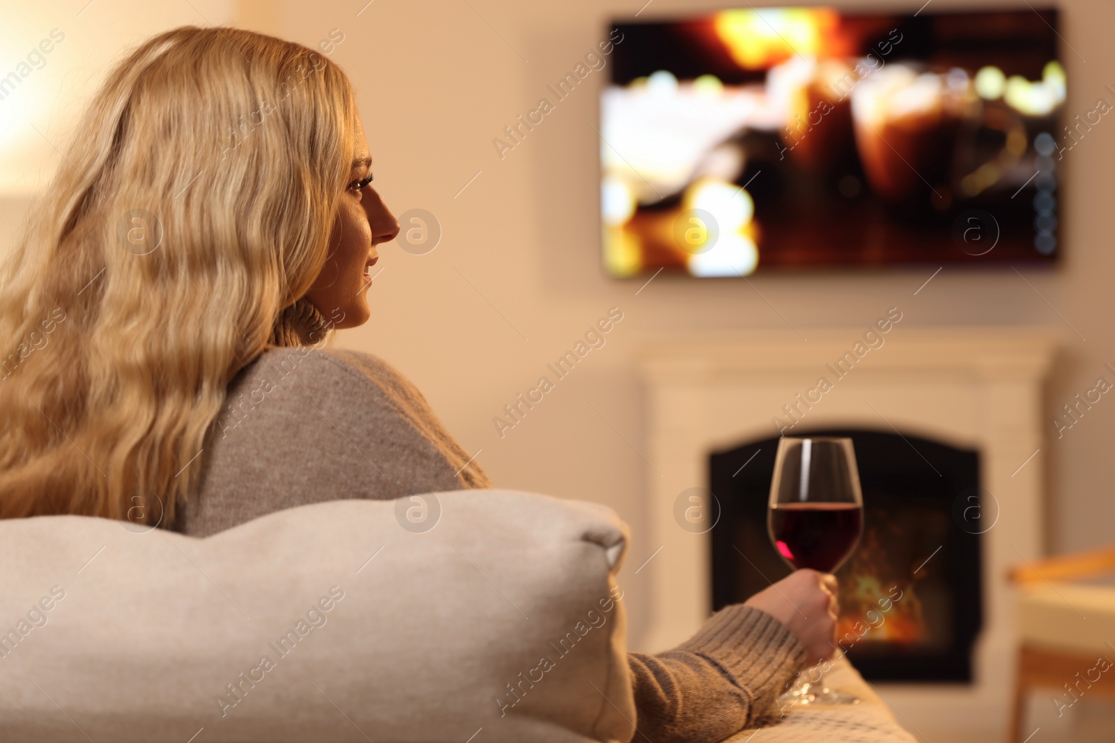 Photo of Beautiful young woman with glass of wine resting near fireplace at home