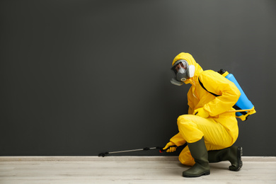 Pest control worker in protective suit spraying pesticide near black wall indoors. Space for text