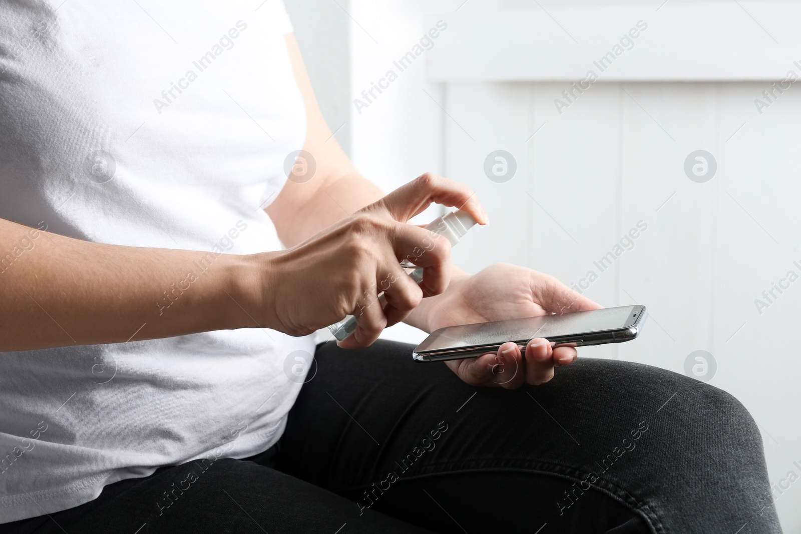 Photo of Woman spraying antiseptic onto smartphone indoors, closeup