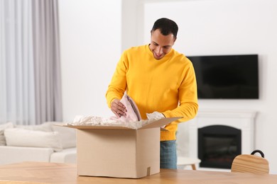 Photo of Happy young man opening parcel at table indoors. Internet shopping