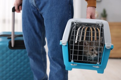 Photo of Travel with pet. Man holding carrier with cute cat and suitcase at home, closeup