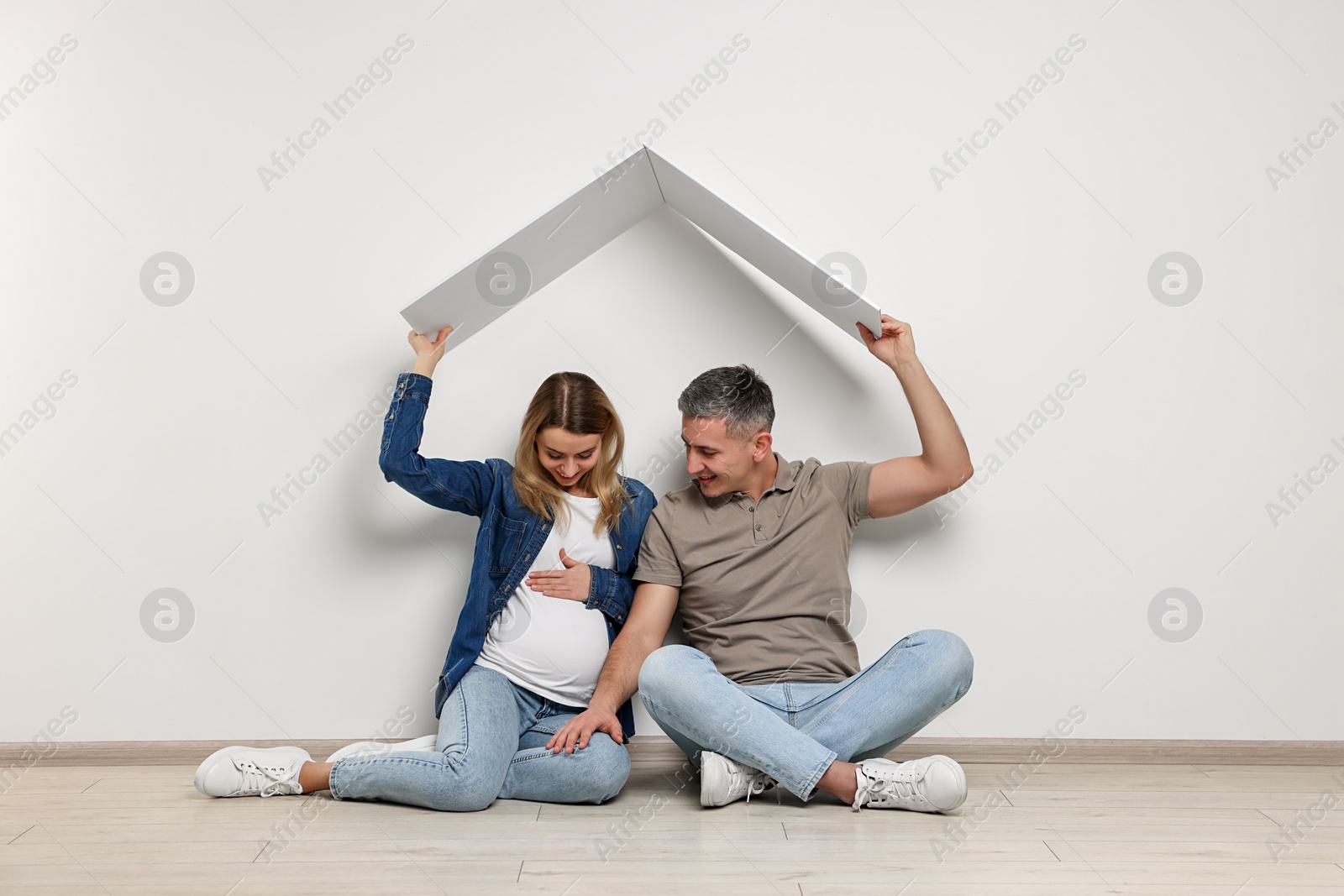 Photo of Young family housing concept. Pregnant woman with her husband sitting under cardboard roof on floor indoors