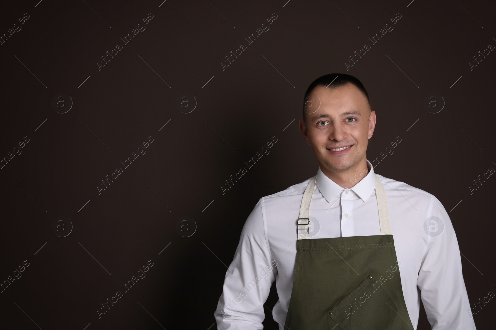 Photo of Portrait of happy young waiter in uniform on brown background, space for text