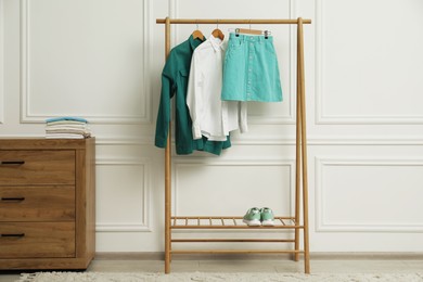 Wardrobe organization. Rack with different stylish clothes, shoes and chest of drawers near white wall indoors