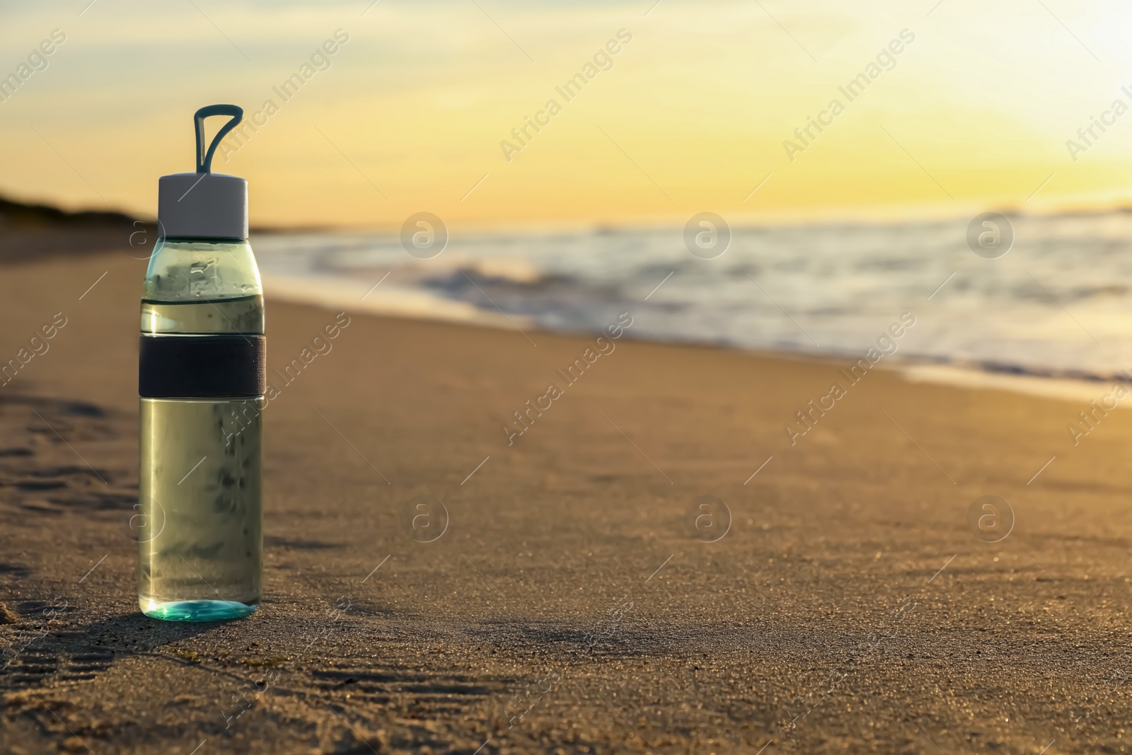 Photo of Glass bottle with water on wet sand near sea at sunset. Space for text