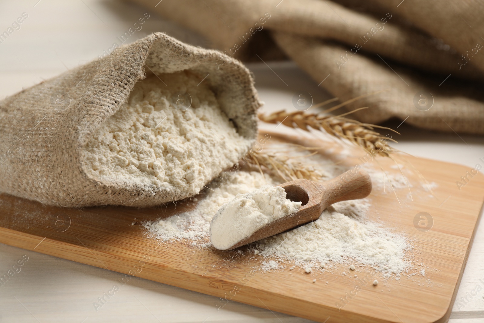 Photo of Sack and scoop of flour with wheat ears on wooden board, closeup