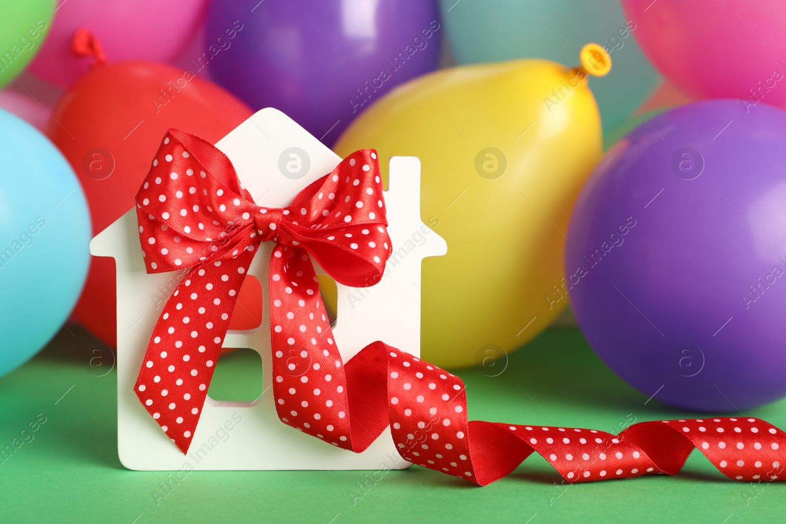 Photo of Wooden house model with red bow and balloons on green background, space for text. Housewarming party