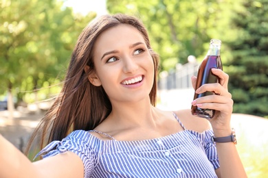 Young woman with bottle of cola taking selfie outdoors