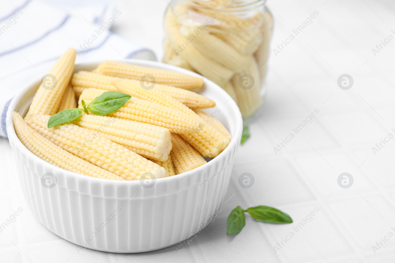 Photo of Canned baby corns with basil on white tiled table, closeup. Space for text