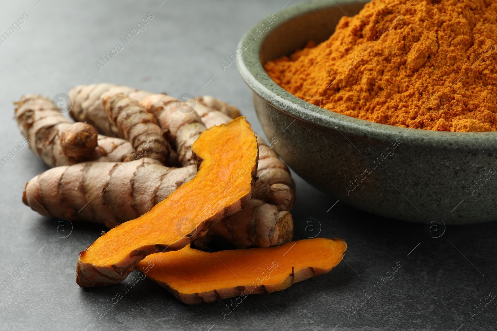 Photo of Bowl with turmeric powder and raw roots on grey table, closeup