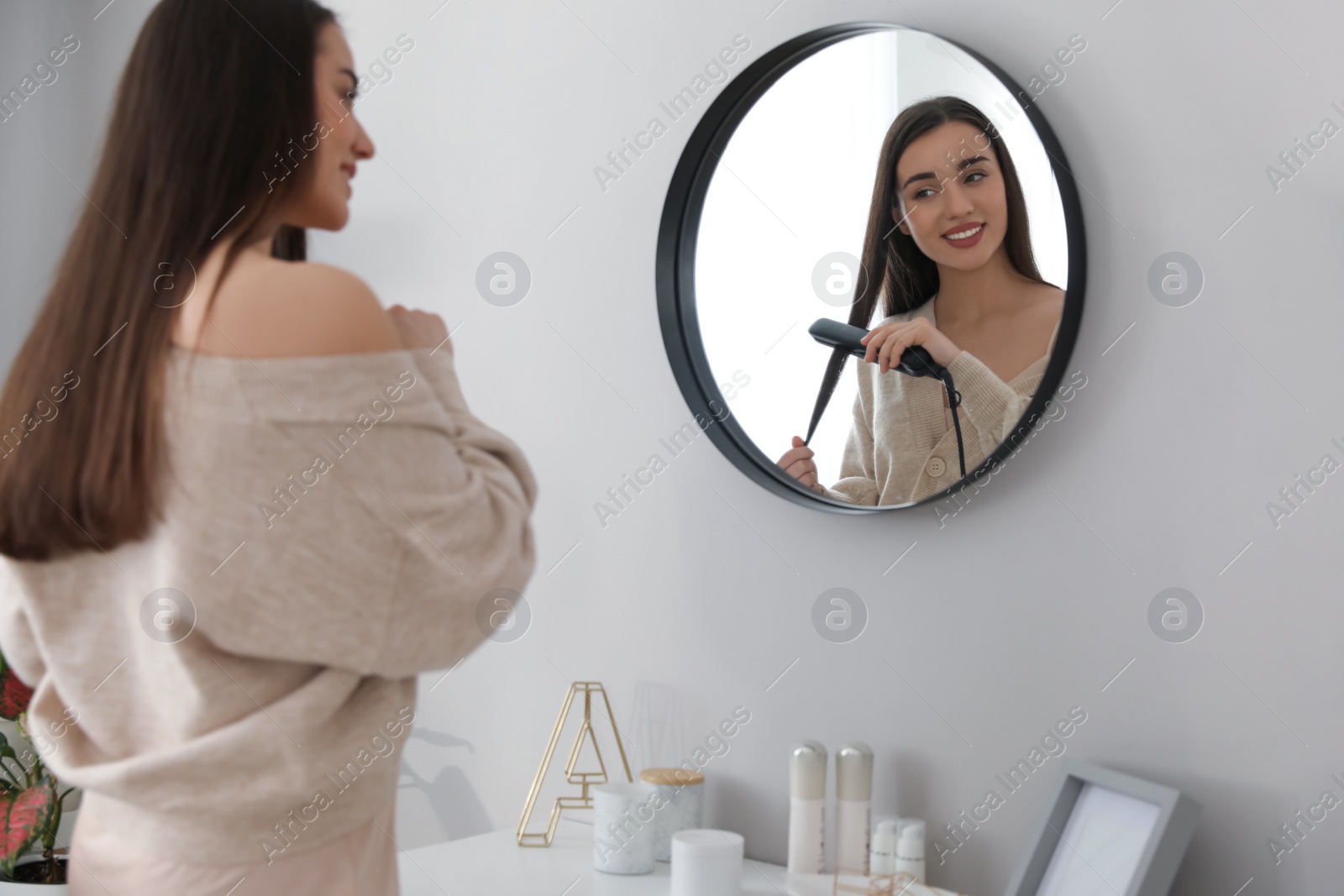 Photo of Young woman straightening hair near mirror at home. Morning routine