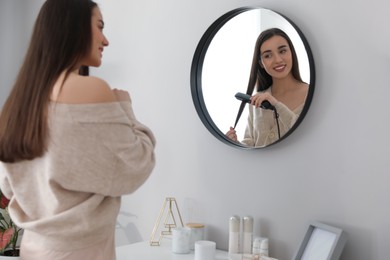 Photo of Young woman straightening hair near mirror at home. Morning routine