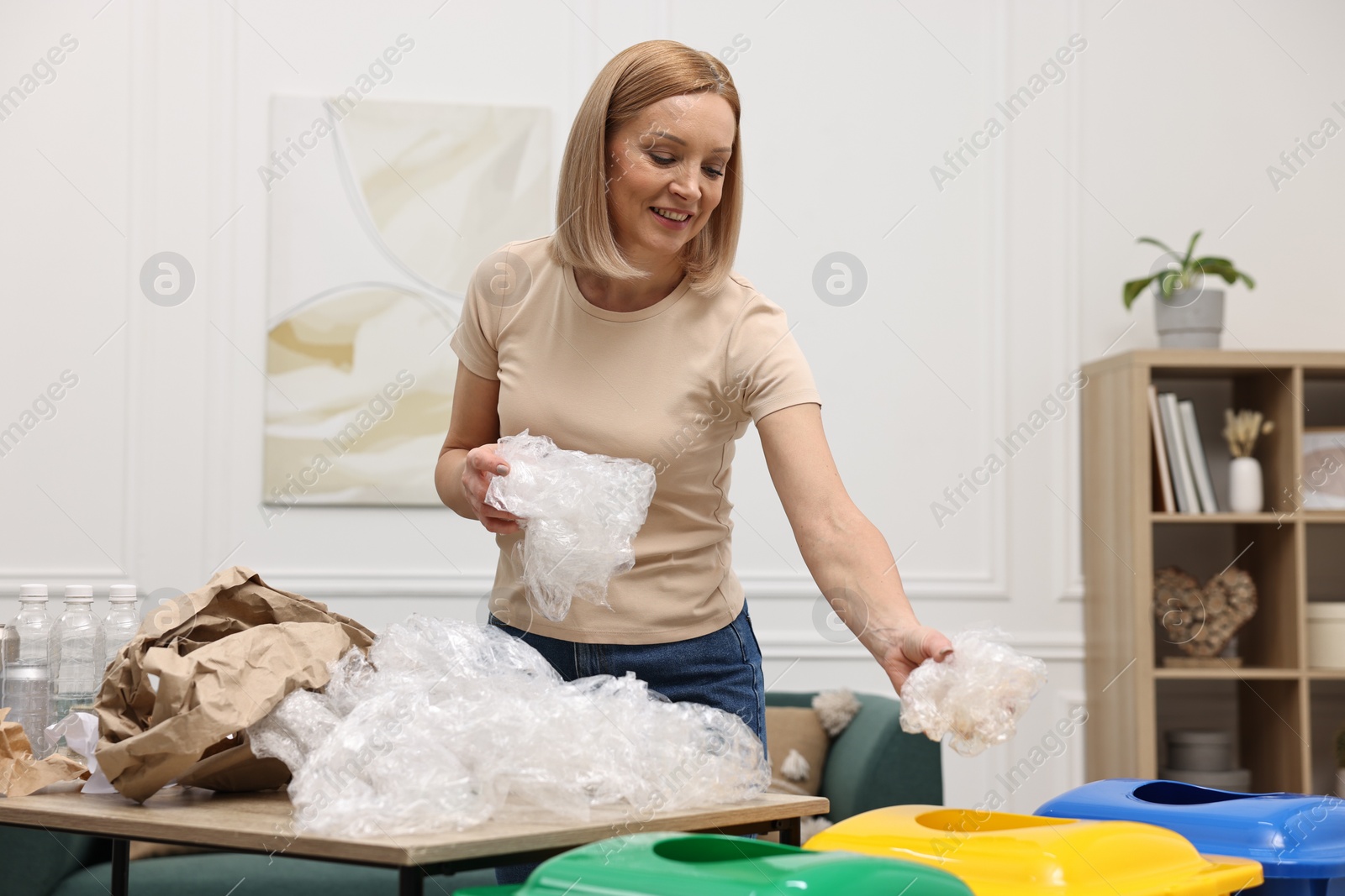 Photo of Garbage sorting. Smiling woman throwing plastic package into trash bin in room