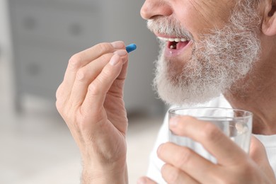 Senior man with glass of water taking pill on blurred background, closeup