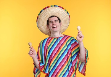 Young man in Mexican sombrero hat and poncho with maracas on yellow background