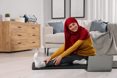 Photo of Muslim woman in hijab stretching near laptop on fitness mat at home. Space for text