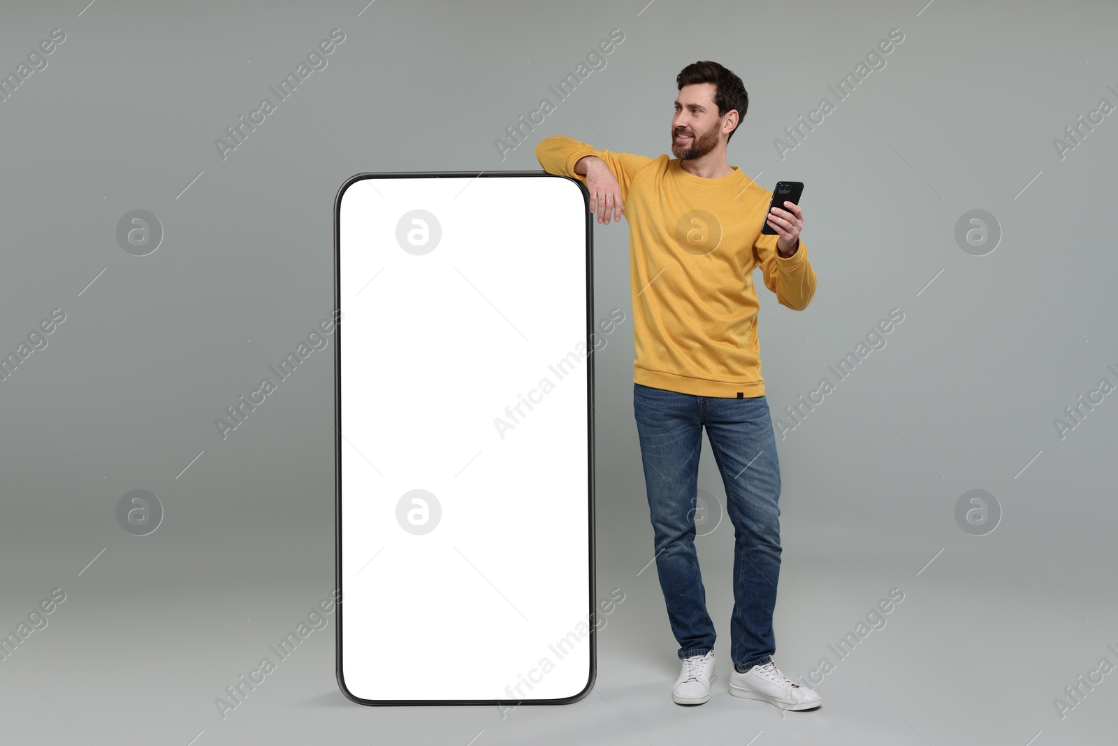 Image of Man with mobile phone standing near huge device with empty screen on grey background. Mockup for design