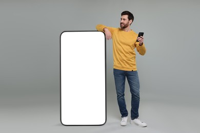 Man with mobile phone standing near huge device with empty screen on grey background. Mockup for design