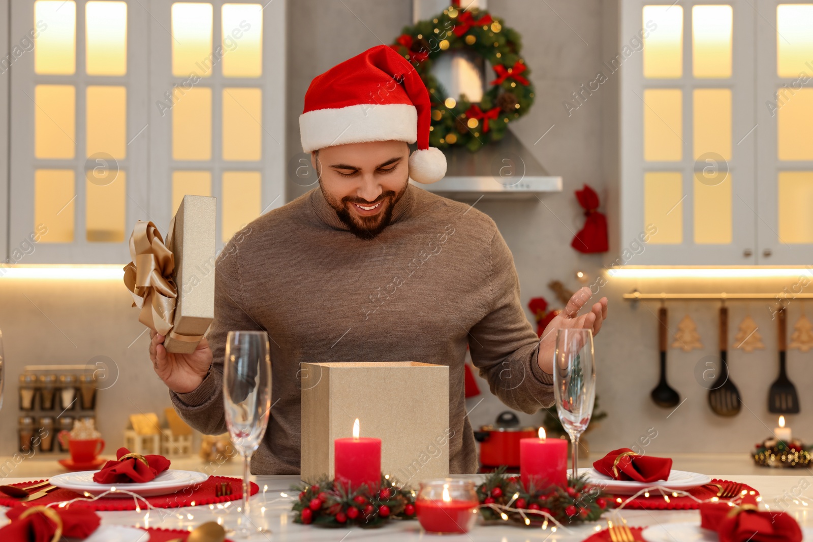 Photo of Happy young man in Santa hat opening Christmas gift at table in kitchen