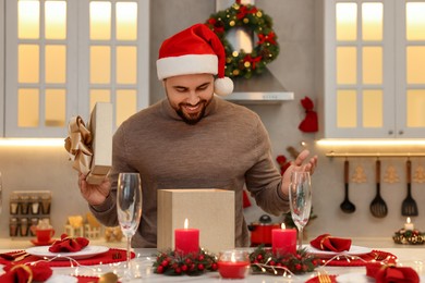 Photo of Happy young man in Santa hat opening Christmas gift at table in kitchen