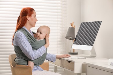 Mother using computer while holding her child in sling (baby carrier) at workplace