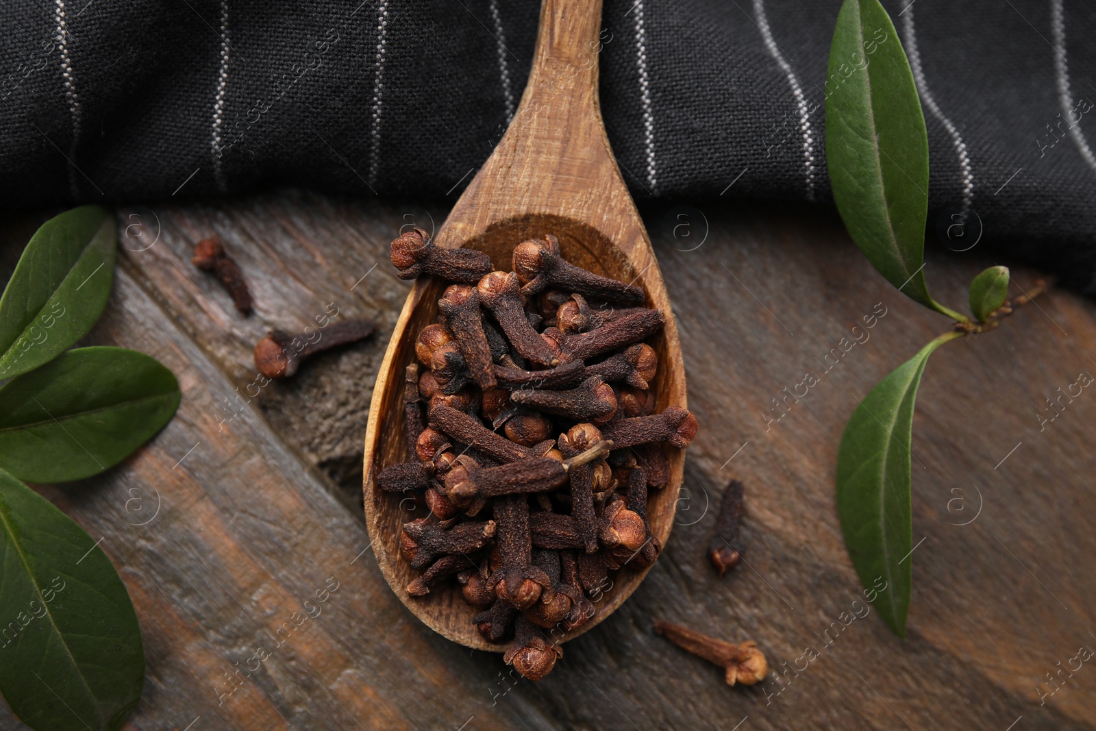 Photo of Spoon with aromatic cloves and green leaves on wooden table, flat lay