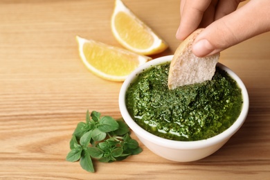 Woman dipping bread into bowl with homemade basil pesto sauce on table