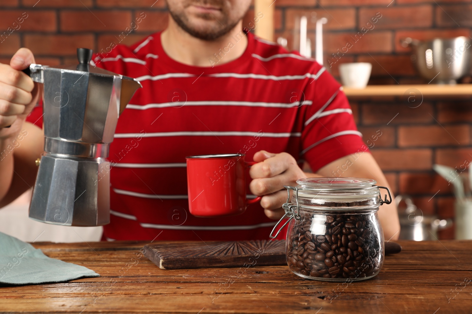 Photo of Brewing coffee. Man with jar of beans, moka pot and mug at wooden table indoors, closeup